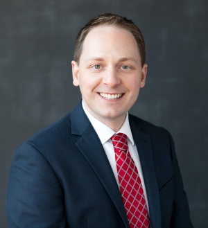 Headshot of Dustin Evatt-Young: man in a blue suit with a white shirt and red tie, smiling
