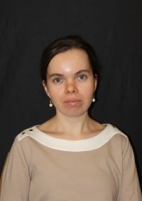 Headshot of Lana Kargetta with brown hair and a beige blouse in front of a black background. 